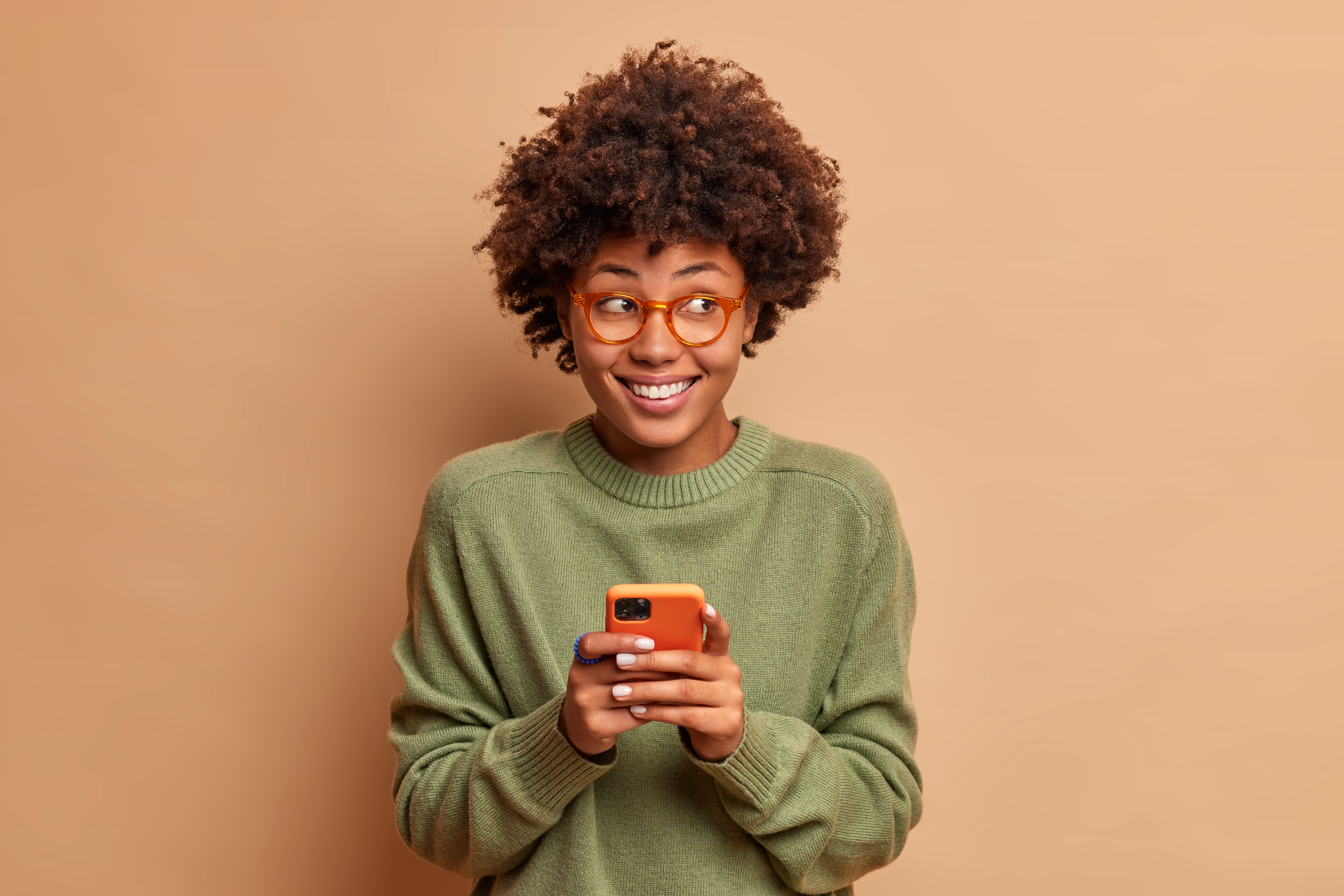 Portrait of good looking woman uses mobile phone receives pleasant feedback in the workplace smiles broadly and looks thoughtfully aside isolated on beige wall