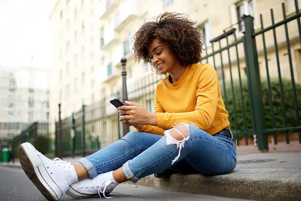 cool young african american woman sitting outside on street with mobile device